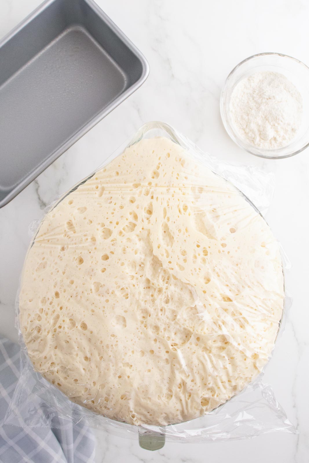 Mashed potato bread dough doubled in size and covered in plastic in a mixing bowl.