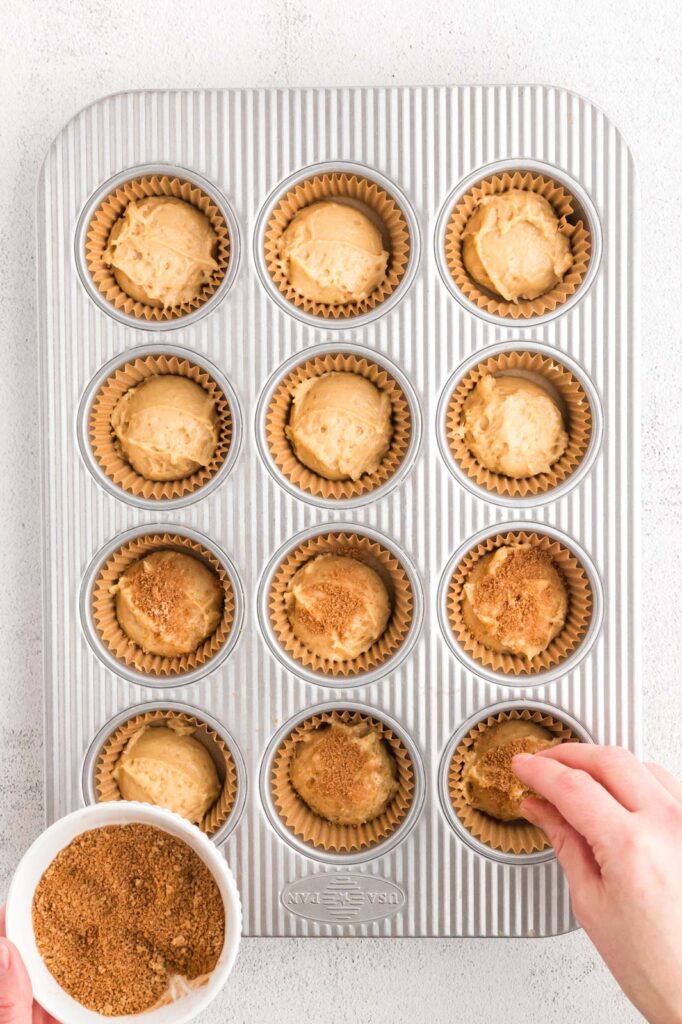 a muffin tin partly full and a hand sprinkling on cinnamon sugar 